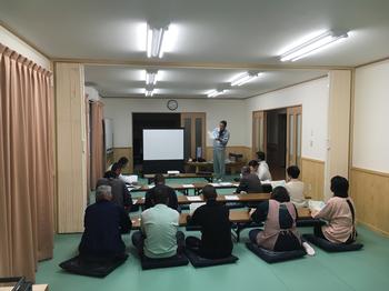 部屋に長い机が並べられ、その前に座布団が敷かれて参加者たちが座り前方に立つ講師の話を聞いている様子を後方から写した写真