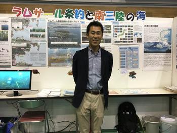 屋内の展示会場で、「ラムサール条約と南三陸の海」と書かれたボードを背景に、ネイビーのジャケットとベージュのパンツを着用したタクゾー博士がが笑顔で立っている写真