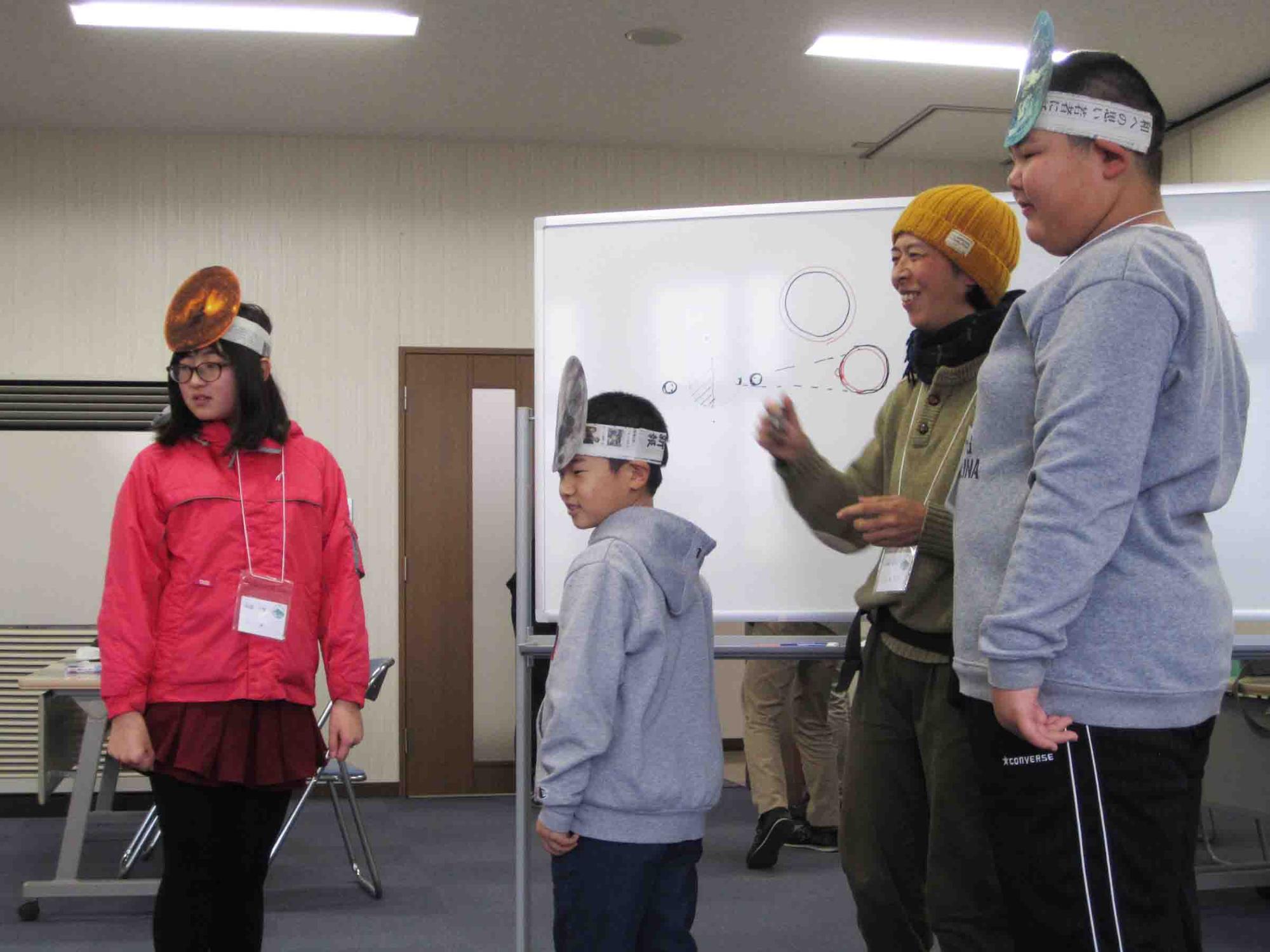 山崎講師と太陽・月・地球の被り物をした3人の子どもたちがホワイトボード前に立っている写真