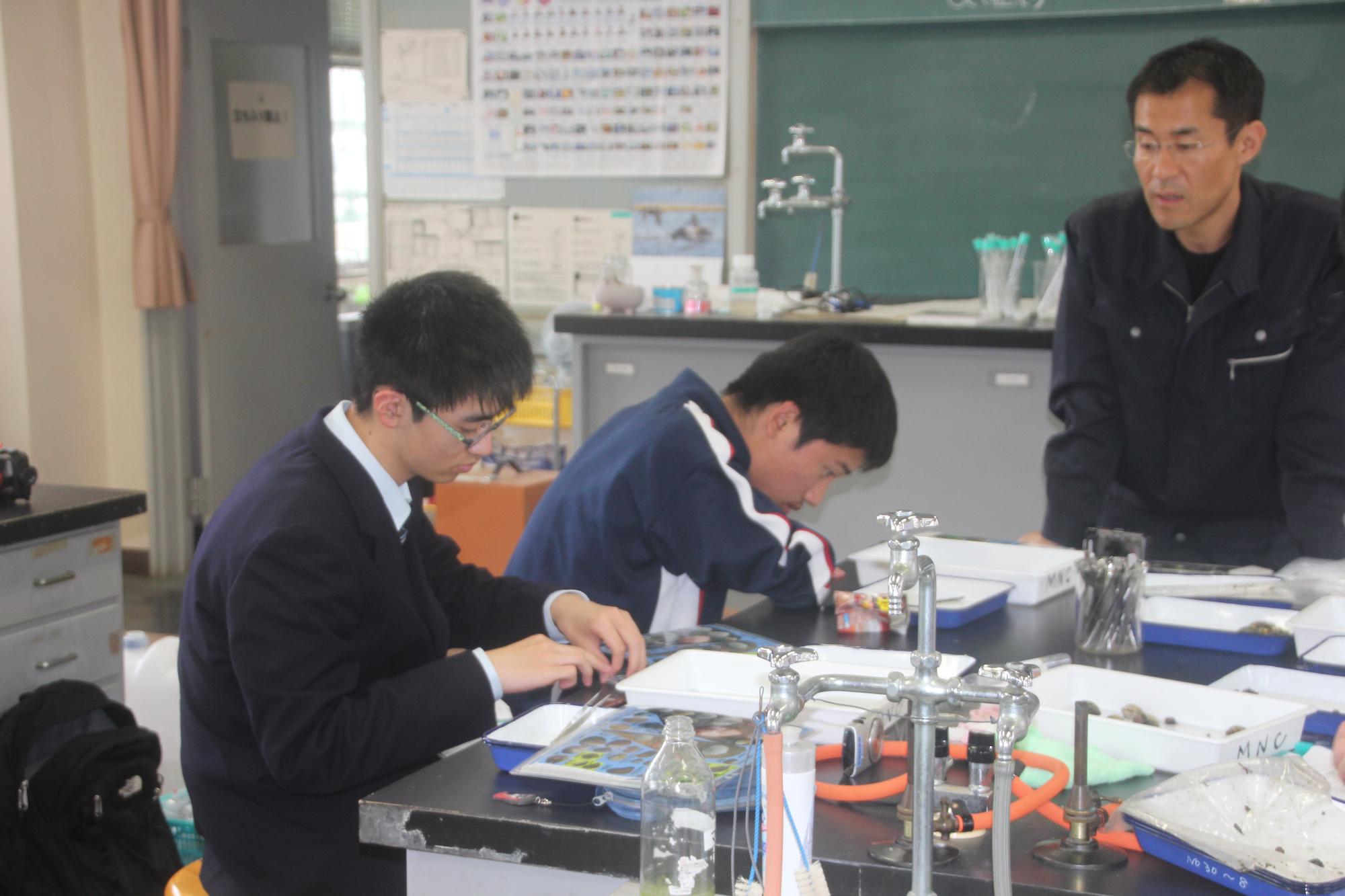 紺色の作業服を着た男性が見ているなか、椅子に座った2名の男子生徒が調査を行っている様子の写真