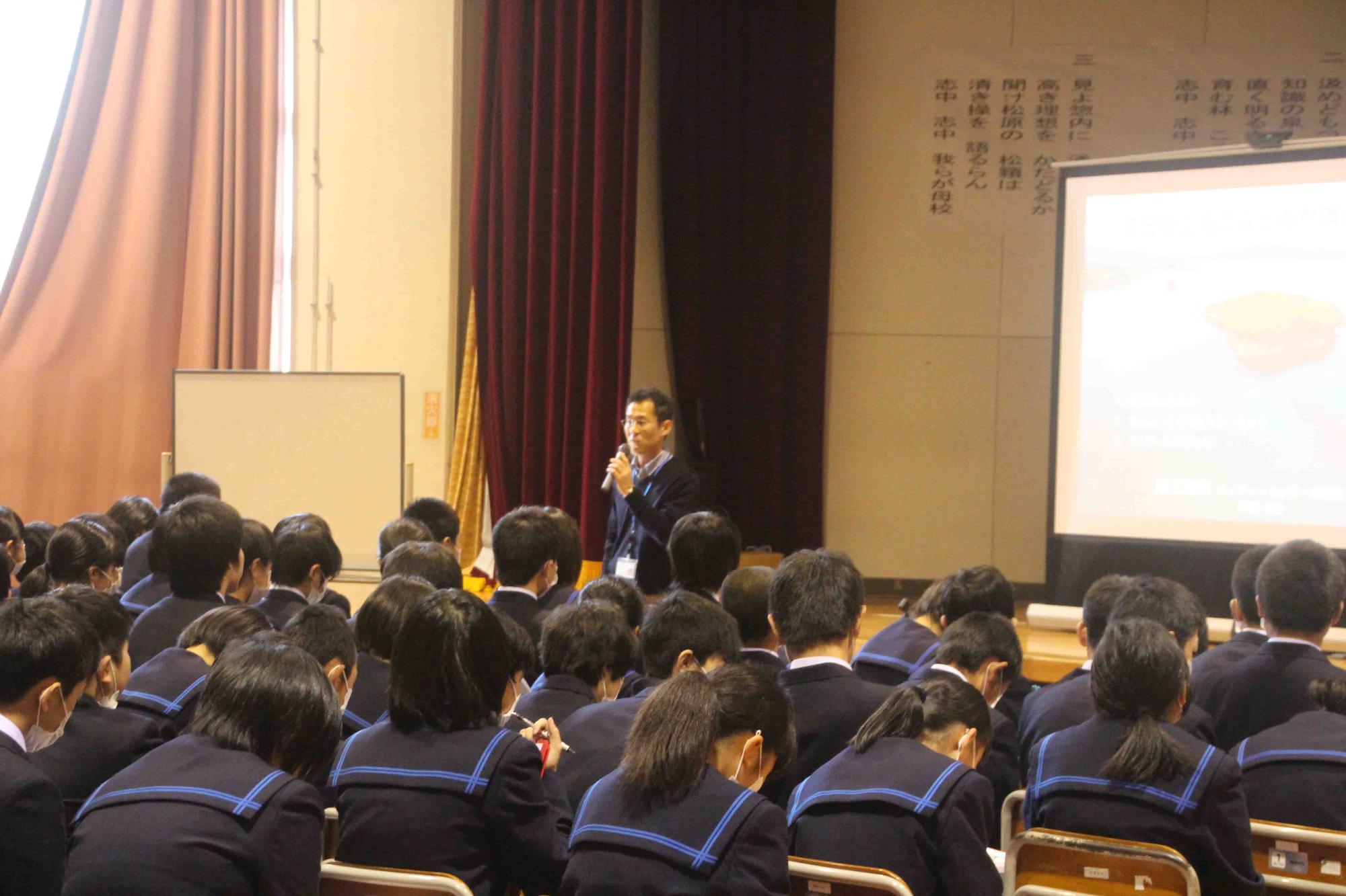 資料が映し出されたスクリーンが設置されたステージの前でマイクを持っている男性の話を、椅子に座った生徒たちが聞いている写真