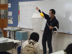 室内のホワイトボードの前に立つ男性が、大きな観察用ネットを持ち使い方を説明している写真