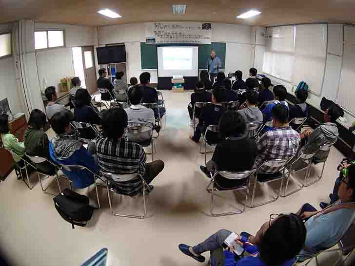 黒板の前に設置されたスクリーンの右側に男性が立ち、パイプ椅子に座った参加者たちが熱心に話を聞いている様子を後方から撮影した写真