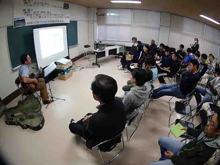 スクリーンの左側に座ってギター演奏を行っている金谷博士を、参加者たちが聞いている写真