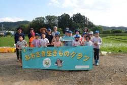 田んぼの前で「おおさき生きものクラブ」と書かれた横断幕を持って集合写真を撮っている子どもたちの写真