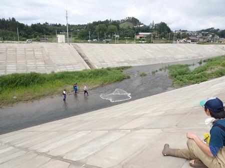 コンクリートで固められた土手の上に座った参加者が、3人の男性が川に入って網を放り投げている様子を見ている写真