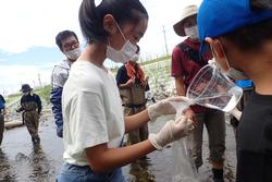 川でマスクと手袋をつけて容器に入った水を袋に移し替えている子どもの写真