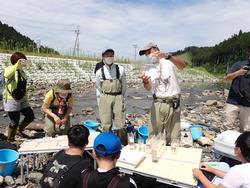 川辺で男性講師が透明な容器を持って説明をしている様子の写真