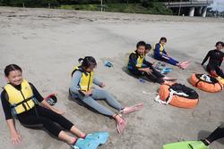 砂浜でウェットスーツを着て足にフィンを履いた子どもたちが一列に並んで座っている写真