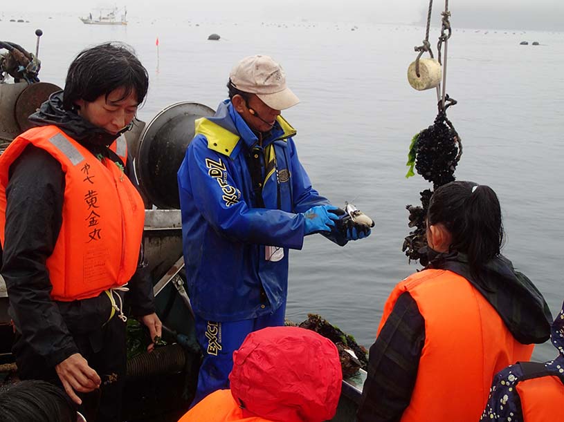 漁船の上でカキを持った男性の話をオレンジ色の救命胴衣を着た子供たちが聞いている写真