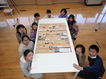 白いテーブルの上に置かれた大きな壁新聞を囲んで集合写真を撮る子どもたちの写真