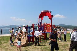 赤い遊具で子どもたちが遊び、大人たちが見守っている海辺の公園の写真