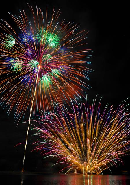 夜空を背景に、水面に反射した赤、オレンジ、緑、青など多色の線香花火のような光を放つ大きな花火と、下部で金色に激しく炸裂する花火を映した写真