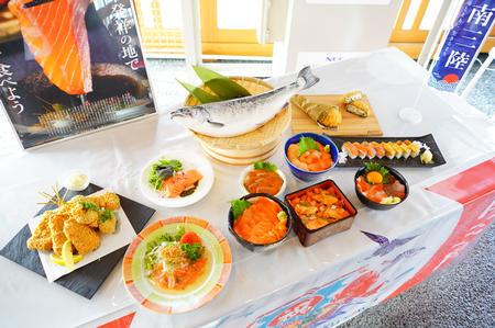 サーモンのポスターの前に置かれた台の上に、ざるに乗ったサーモンと、サーモンを使った天ぷらやすし、海鮮丼など様々な料理が並んでいる写真